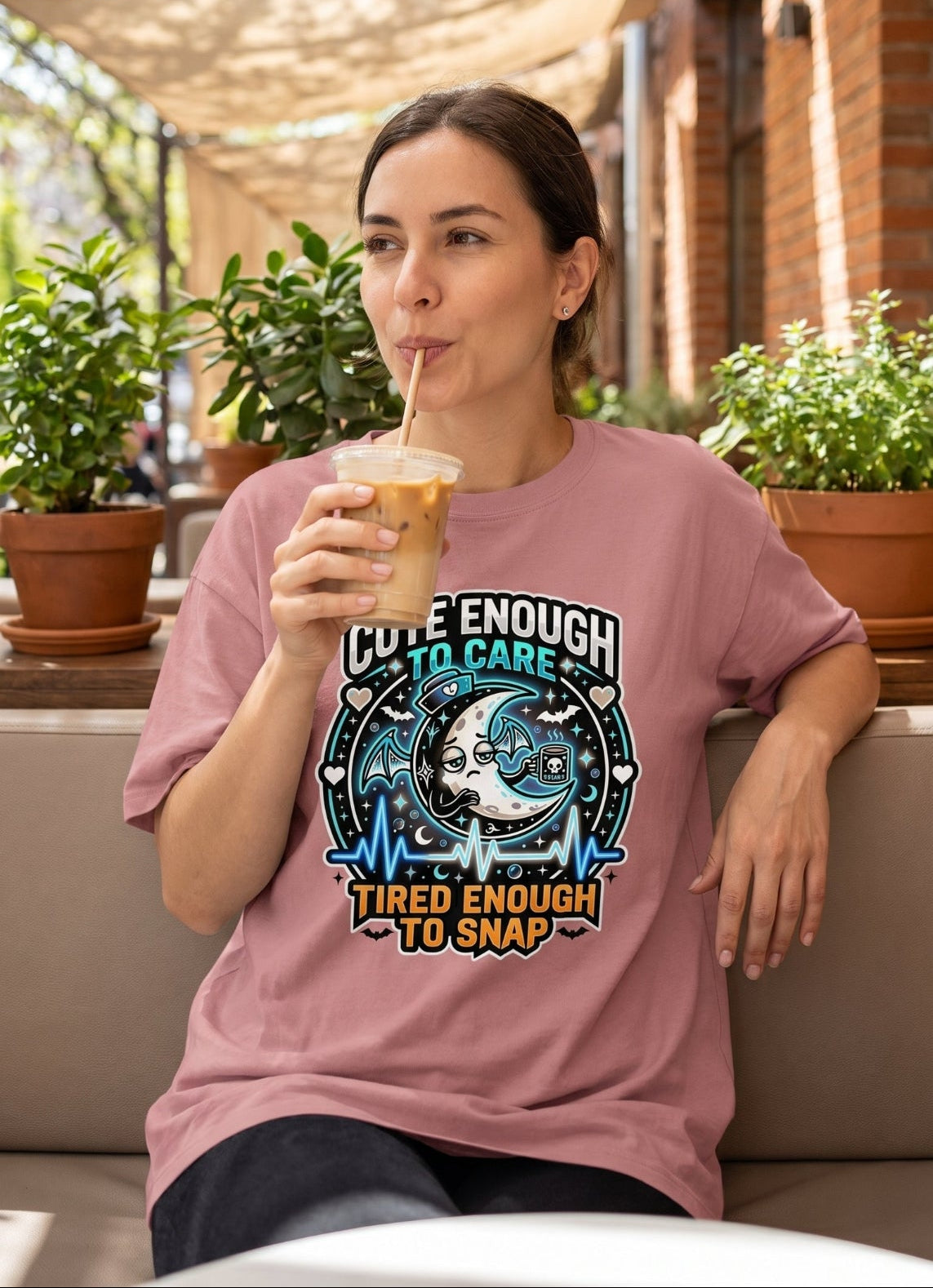 A woman sitting at an outdoor cafe wearing a dusty pink graphic t-shirt with a blue and white design that reads 'CUTE ENOUGH TO CARE TIED ENOUGH TO SNAP'. She is holding an iced coffee.
