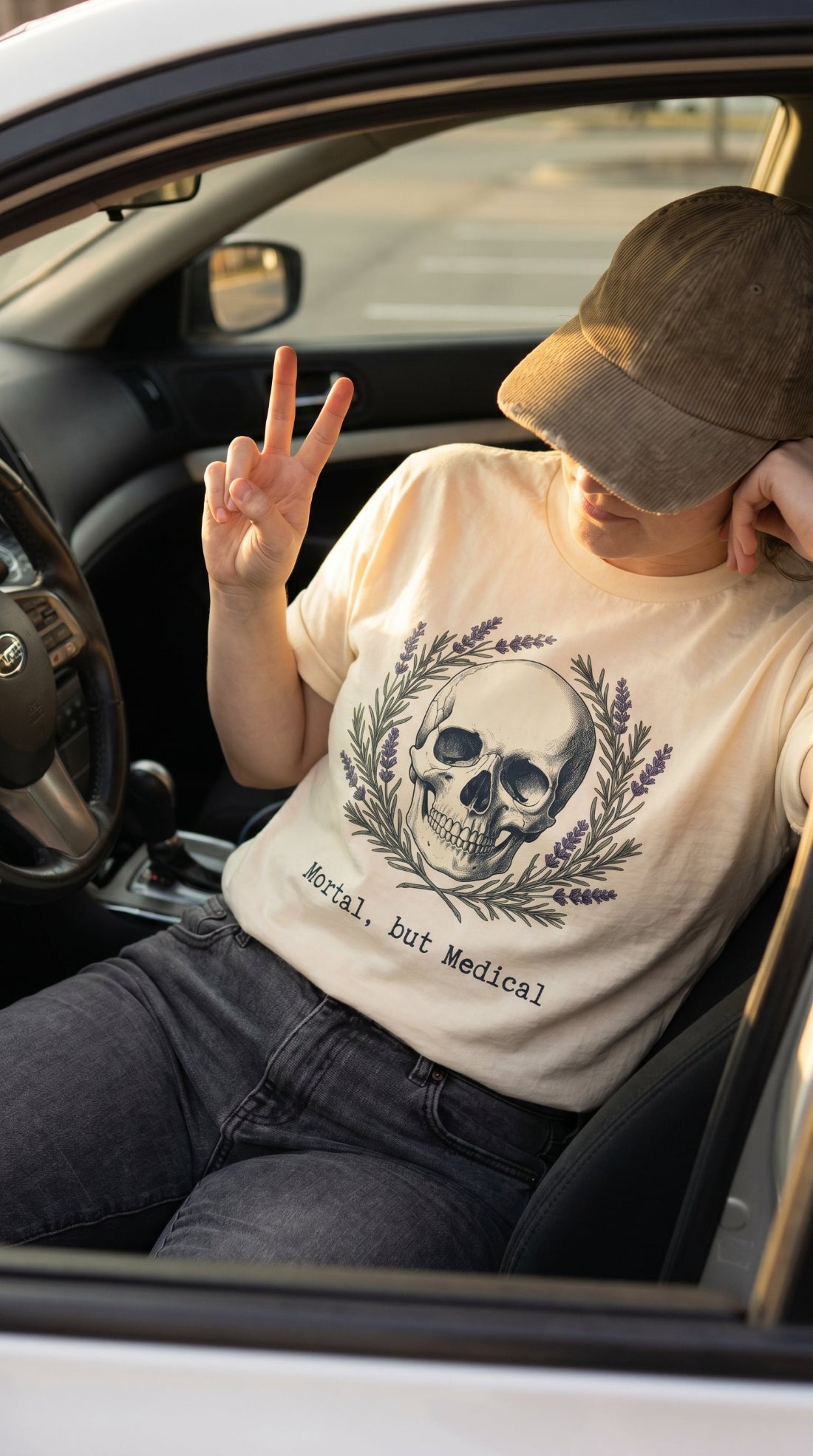 A person sitting in a car wearing a cream graphic t-shirt with a skull and wreath design, dark gray jeans, and a brown baseball cap, making a peace sign.